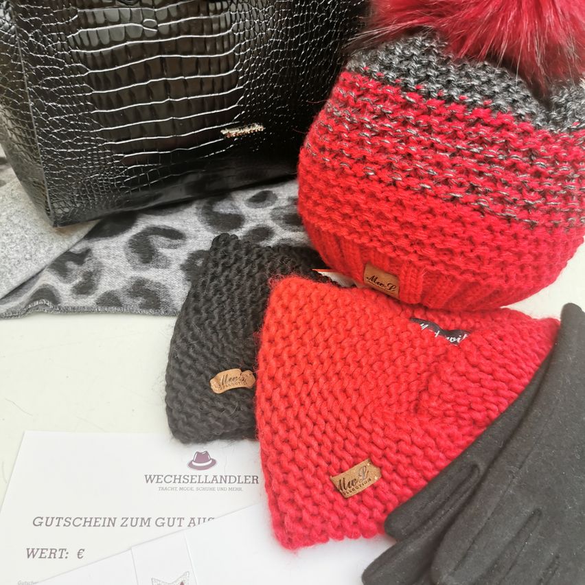 Eine rote gestrickte Mütze, eine graue Mütze und Handschuhe sind auf einer weißen Oberfläche ausgestellt. Ein Leopardenmusterteppich und eine schwarze Lederhandtasche sind im Hintergrund. Ein Gutschein mit 'Gutschein zum Gut Aus' ist vorhanden.