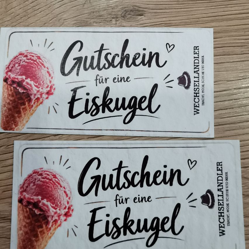 Two white cards with black text and an image of a strawberry ice cream cone. The text reads 'Gutschein für eine Eiskugel'. The cards are placed on a wooden surface.