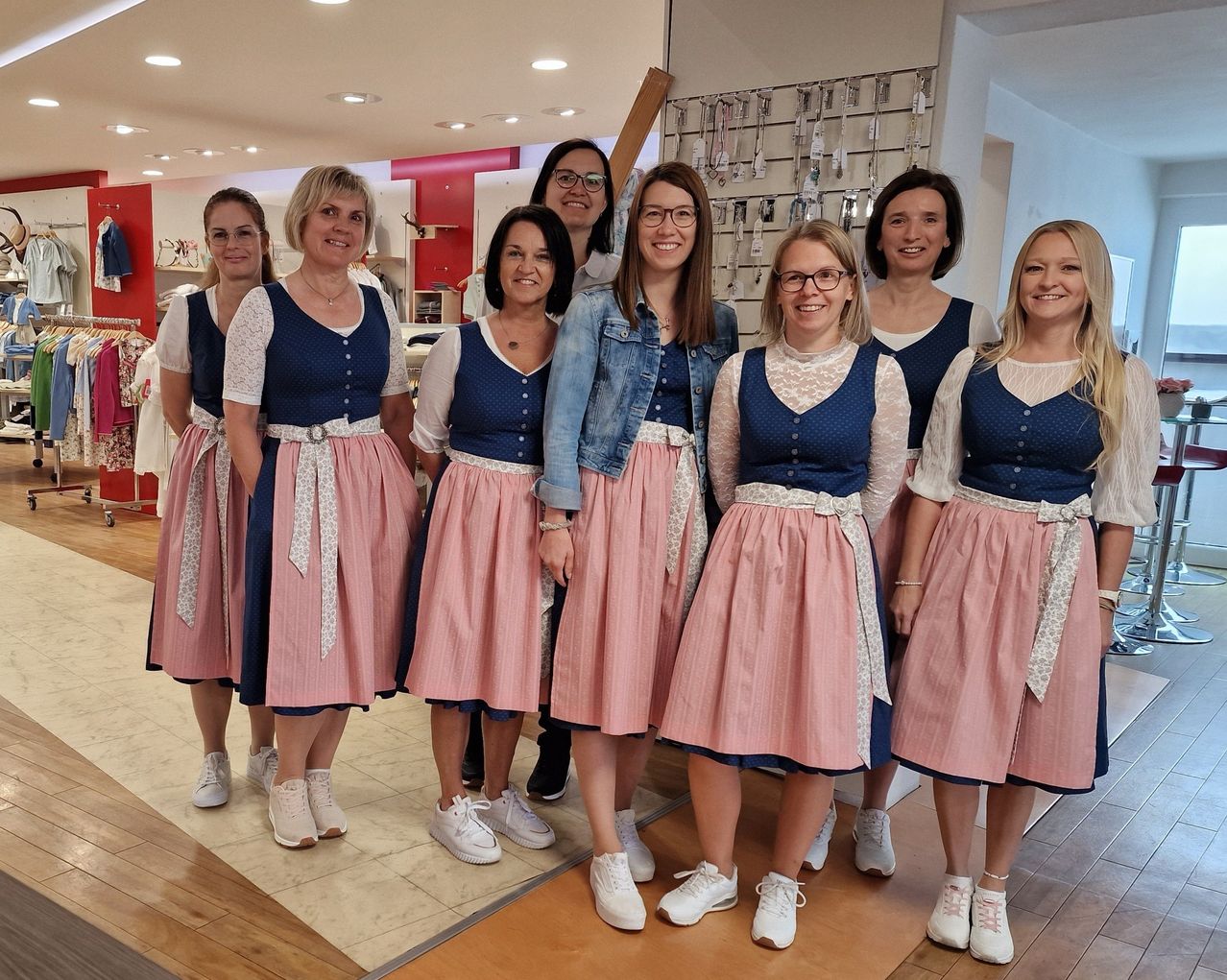Eine Gruppe von Frauen in passenden Kleidern steht in einem Bekleidungsgeschäft und lächelt für ein Foto. Sie tragen verschiedenfarbige Kleider und weiße Sneaker. Ein Holzboden und Deckenleuchten sind sichtbar.