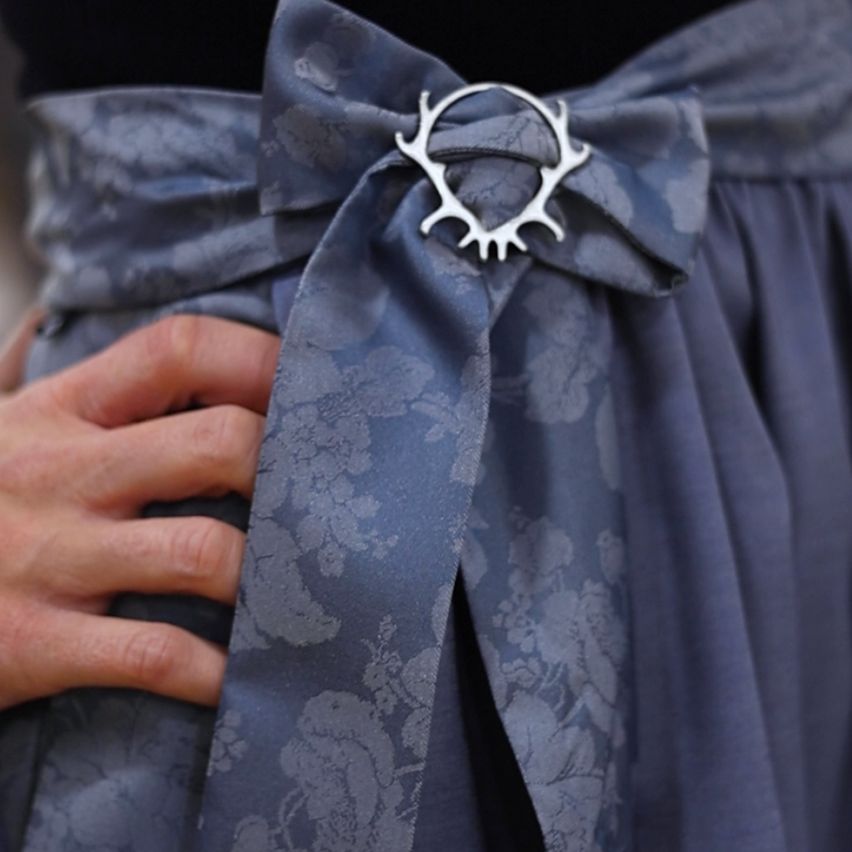 Ein Nahaufnahme von einer Person, die ein blaues Kleid trägt, mit einer silbernen Geweihbrosche an der Taille. Der Stoff hat ein Blumenmuster.