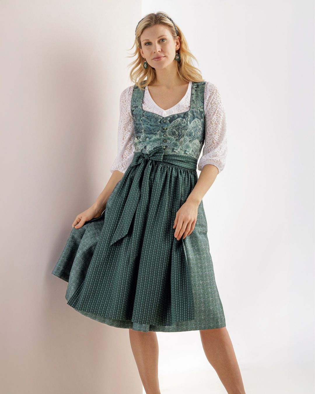 Eine Frau mit blonden Haaren steht an einer weißen Wand und trägt ein grünes Kleid mit einer weißen Spitzenbluse und einem weißen Gürtel. Sie posiert für ein Foto.