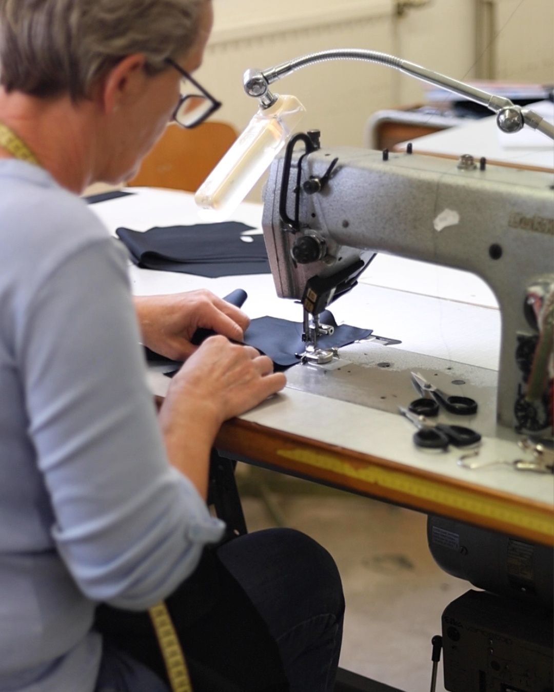 An individual wearing glasses is sitting and using a sewing machine on a white desk, with scissors and other tools nearby.