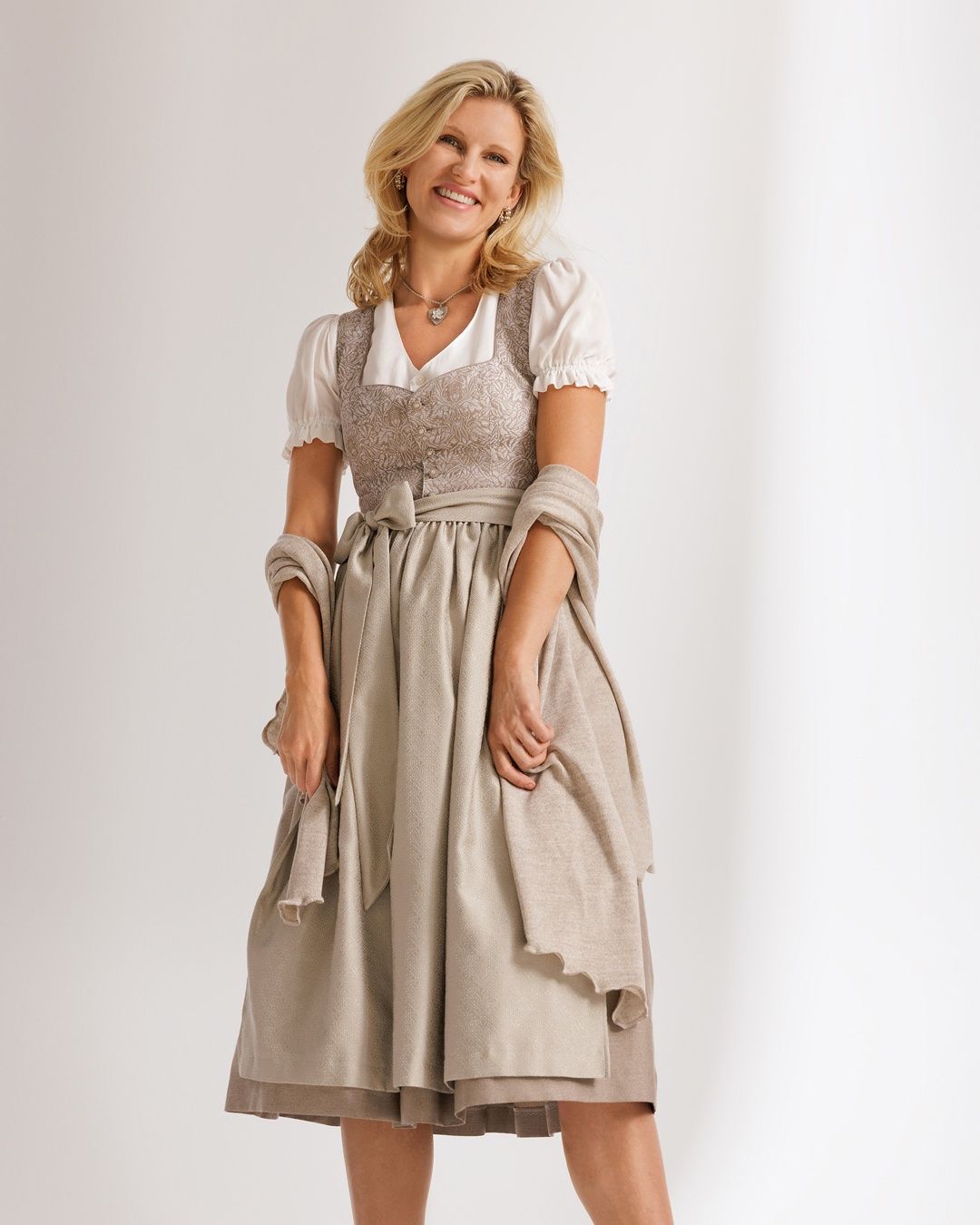 Eine lächelnde Frau in einem traditionellen deutschen Kleid, mit einer Halskette und einem Ring, steht mit den Händen in den Taschen.