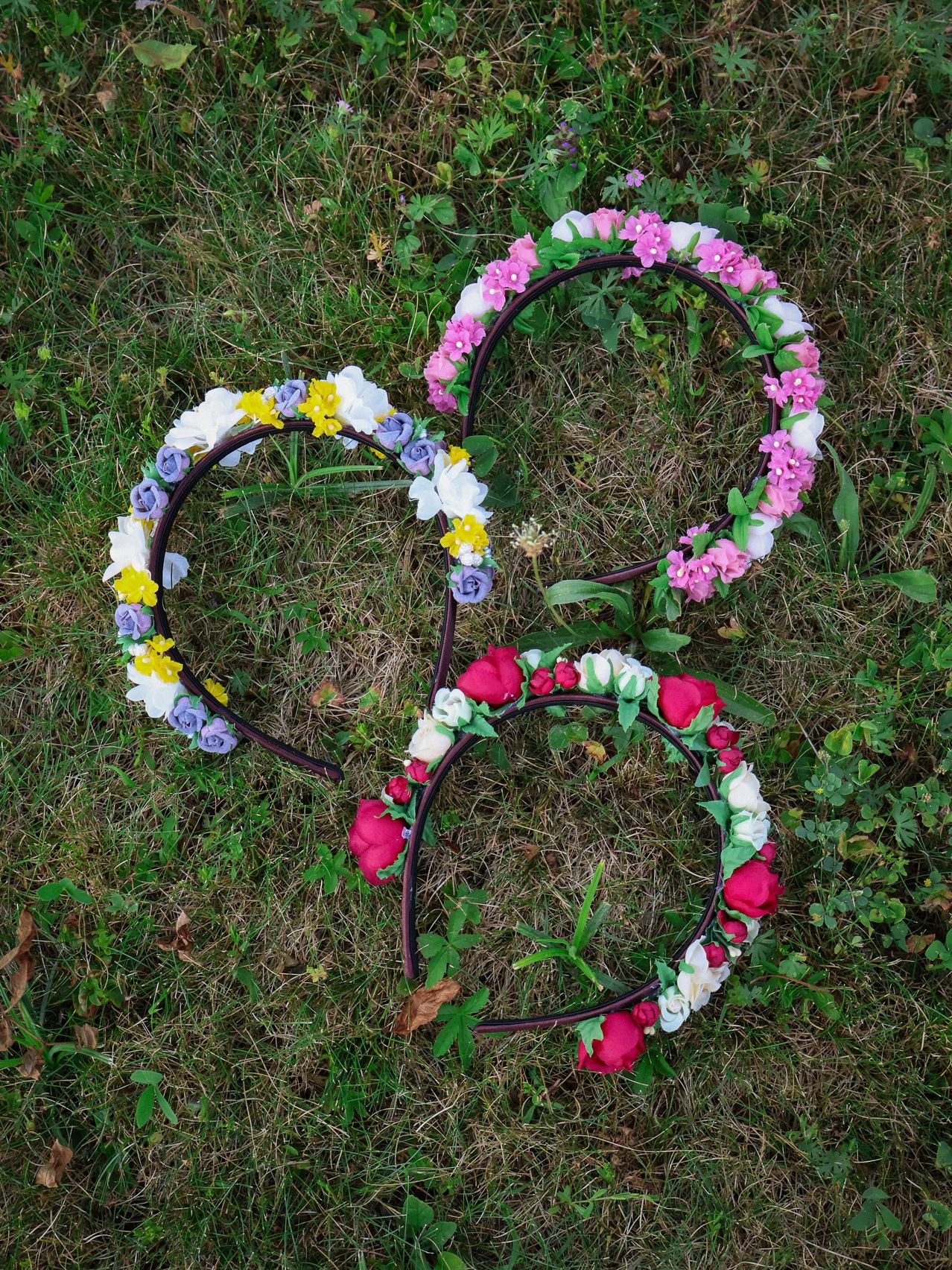 Drei Blumenkränze, jeder mit unterschiedlich farbigen Blumen, liegen auf einer Grasfläche. Der linke Kranz hat weiße, gelbe und lila Blumen. Der mittlere hat rosa und weiße Blumen. Der rechte hat rote, weiße und rosa Blumen.