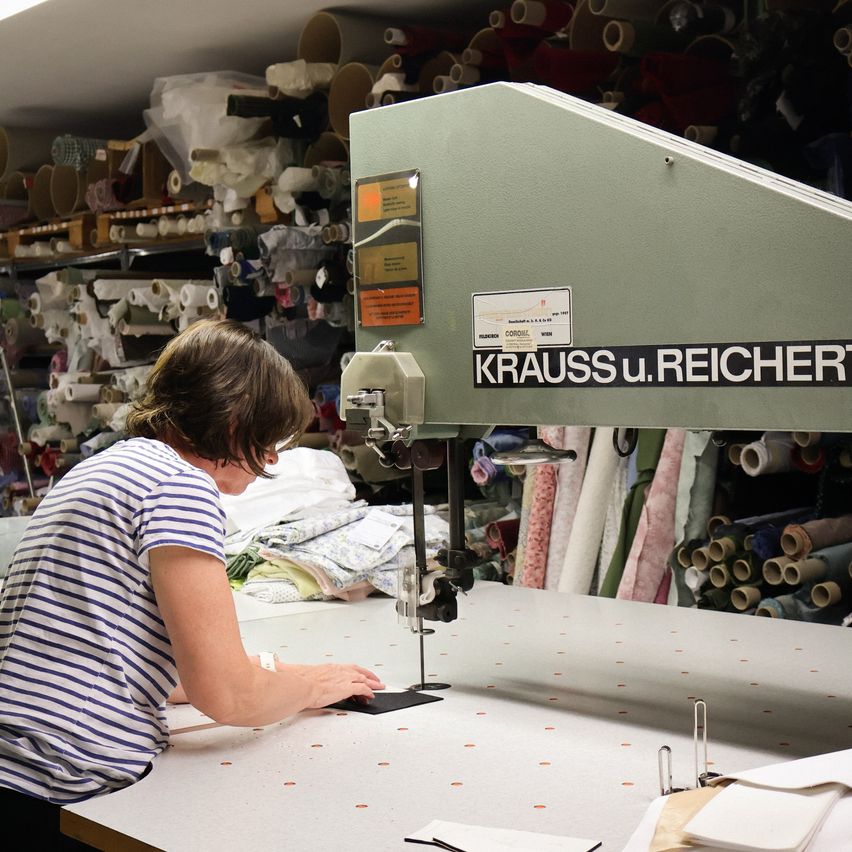 A woman in a striped shirt is operating a large sewing machine labeled 'KRAUSSU.REICHERT' in a textile workshop. The workshop is filled with fabric rolls and sewing equipment.