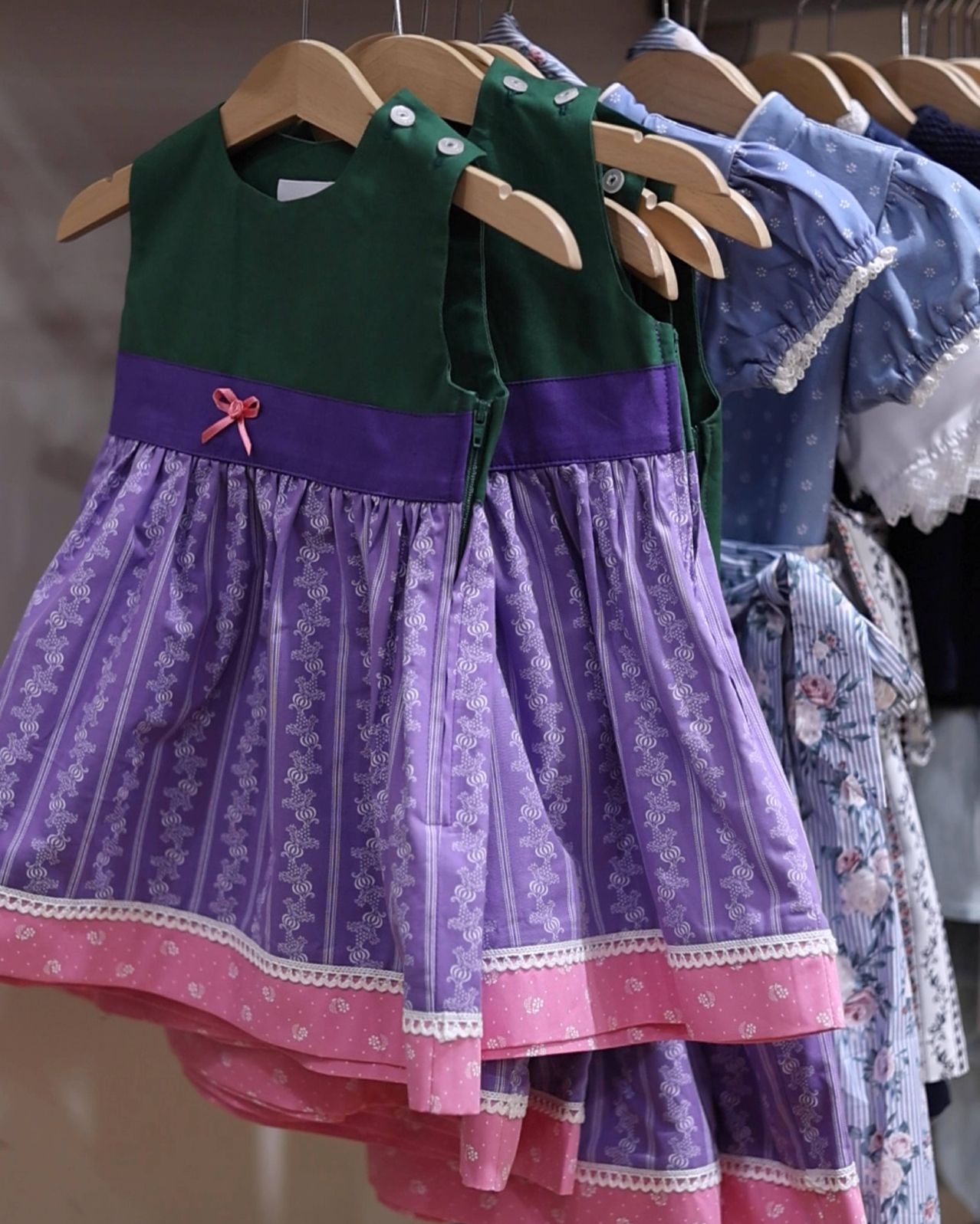 Ein lila-pinkes Kleid mit weißen Bordüren hängt an einem Holzbügel, neben einem anderen Kleid. Das lila Kleid hat ein Blumenmuster und eine pinke Schleife auf der Vorderseite.