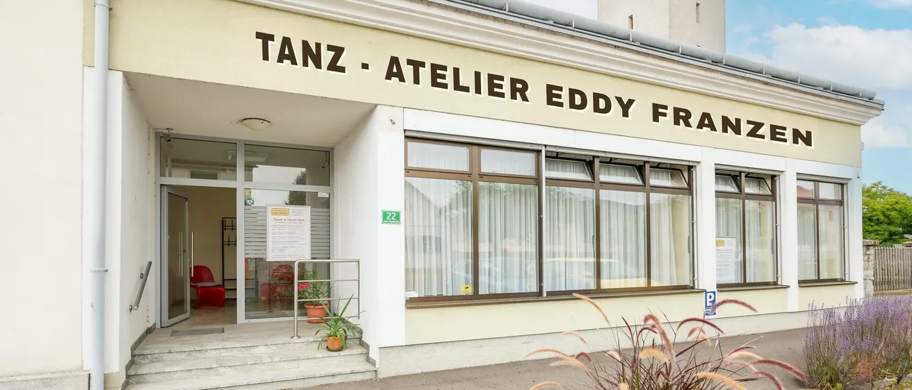 Die Vorderseite eines Gebäudes mit der Aufschrift Tanz Atelier Eddy Franz. Der Eingang hat Glastüren, und es gibt ein Schild mit Text. Es gibt Topfpflanzen und Stühle auf der Veranda. Ein Turm befindet sich auf der rechten Seite.