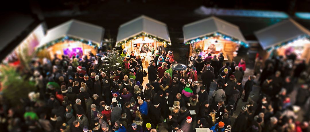 Eine Menschenmenge in Winterkleidung steht in einem Kreis um einige Stände. Die Stände haben Lichter und Dekorationen. Es scheint ein Weihnachtsmarkt zu sein.