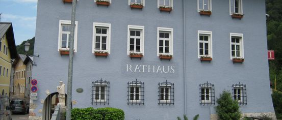 The blue building has the word Rathaus written on it. It has many windows and flower boxes. There is a statue and a street lamp in front.
