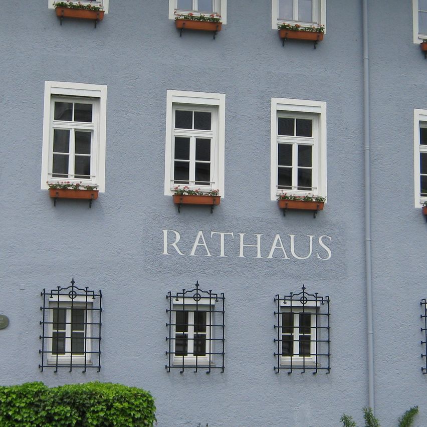Das blaue Gebäude hat das Wort Rathaus darauf geschrieben. Es hat viele Fenster und Blumenkästen. Davor steht eine Statue und eine Straßenlaterne.