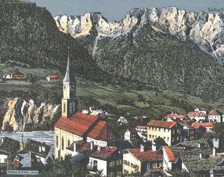 Eine Luftaufnahme eines Dorfes mit rot gedeckten Häusern und einem Kirchturm. Das Dorf ist von üppigen grünen Hügeln und Bergen in der Ferne umgeben.
