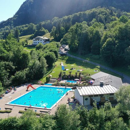 Ein Luftbild eines Resorts mit einem großen Swimmingpool, einem Gebäude und Menschen in der Umgebung. Das Gebäude hat ein graues Dach, und mehrere Autos sind in der Nähe geparkt.