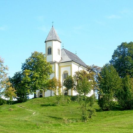 Eine kleine weiße Kirche mit einem grauen Dach und einem Turm steht auf einem grasbewachsenen Hügel, umgeben von Bäumen und Sträuchern unter einem blauen Himmel.