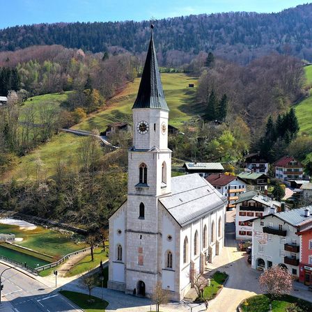 Luftaufnahme einer Kirche mit hohem Turm und Uhr, eingebettet in ein malerisches Dorf, umgeben von üppiger Grünfläche und Bergen.