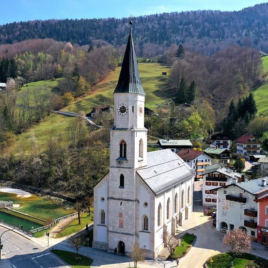 Luftaufnahme einer Kirche mit hohem Turm und Uhr, eingebettet in ein malerisches Dorf, umgeben von üppiger Grünfläche und Bergen.