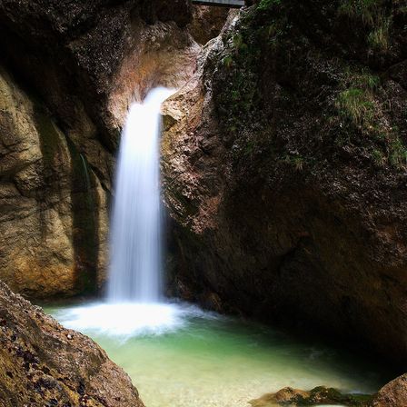 Ein Wasserfall stürzt von einem felsigen Abgrund in einen klaren Pool mit grünem Wasser, umgeben von moosbewachsenen Felsen.