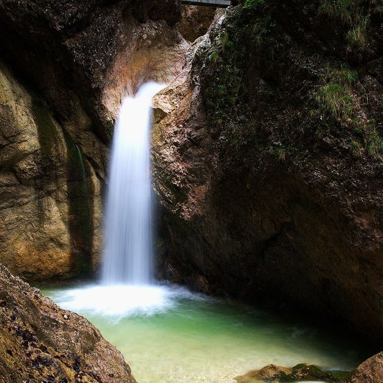 Ein Wasserfall stürzt von einem felsigen Abgrund in einen klaren Pool mit grünem Wasser, umgeben von moosbewachsenen Felsen.