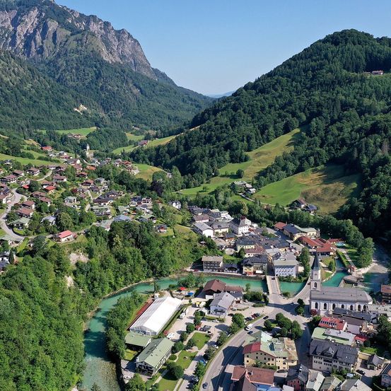 Eine Luftaufnahme eines Dorfes, umgeben von Bergen und Bäumen. Das Dorf liegt in der Nähe eines Flusses und hat Gebäude verschiedener Größen und Farben.