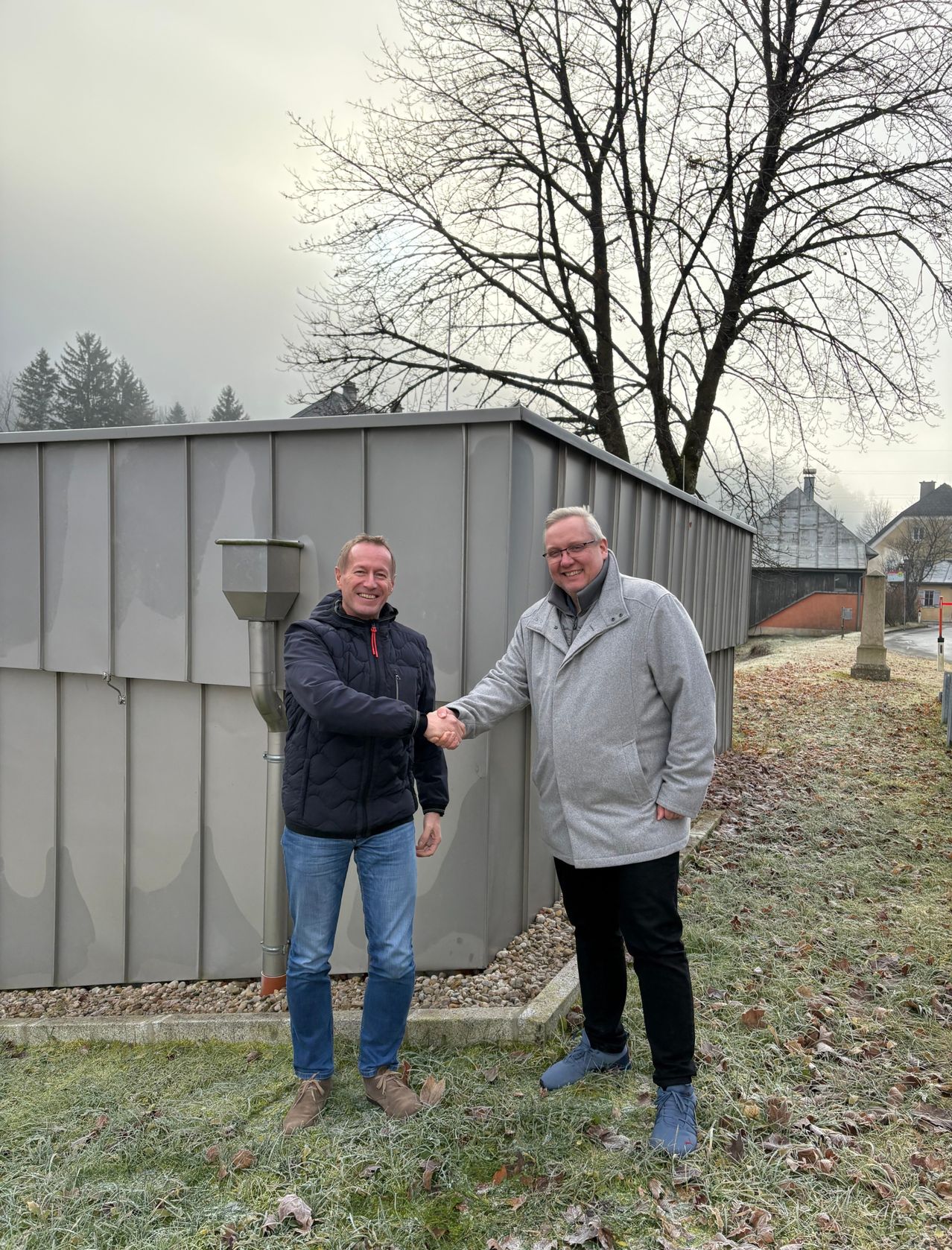 Zwei Männer stehen und schütteln sich die Hände vor einem grauen Gebäude. Sie lächeln beide und scheinen für ein Foto zu posieren. Hinter ihnen steht ein blattloser Baum und ein Weg. In der Ferne befinden sich Bäume und Gebäude.