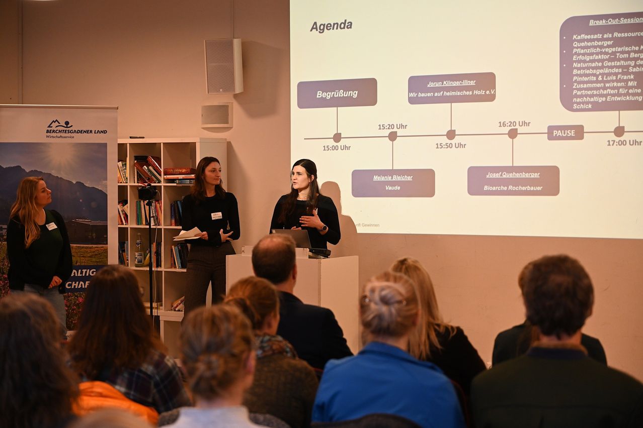 Zwei Frauen stehen vor einer großen Leinwand mit einer Präsentationsagenda, eine hält ein Papier in der Hand, neben einem Mikrofon. Ein Publikum sitzt vor ihnen.