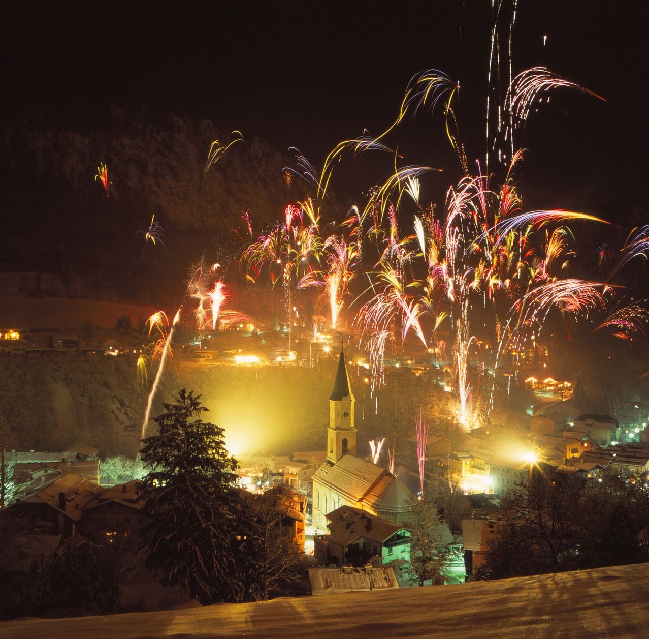 Ein nächtliches Feuerwerk beleuchtet ein Dorf, mit einer Kirche im Zentrum und Gebäuden, die die Landschaft säumen, gegen einen dunklen Himmel.