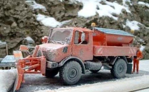 Ein roter Schneepflug auf einer verschneiten Straße mit einer Person in orangefarbener Kleidung, die in der Nähe steht.