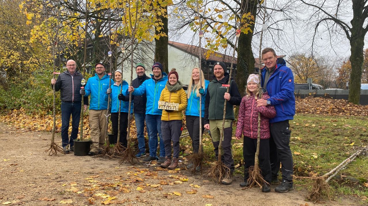 Eine Gruppe von Menschen steht in einem Außenbereich, möglicherweise einem Park, mit Bäumen und gefallenen Blättern. Sie halten alle Stöcke mit Wurzeln und tragen Winterkleidung. Ein Mädchen in einer pinken Jacke lächelt. Der Bereich ist von Bäumen und einem Gebäude mit rotem Dach umgeben.