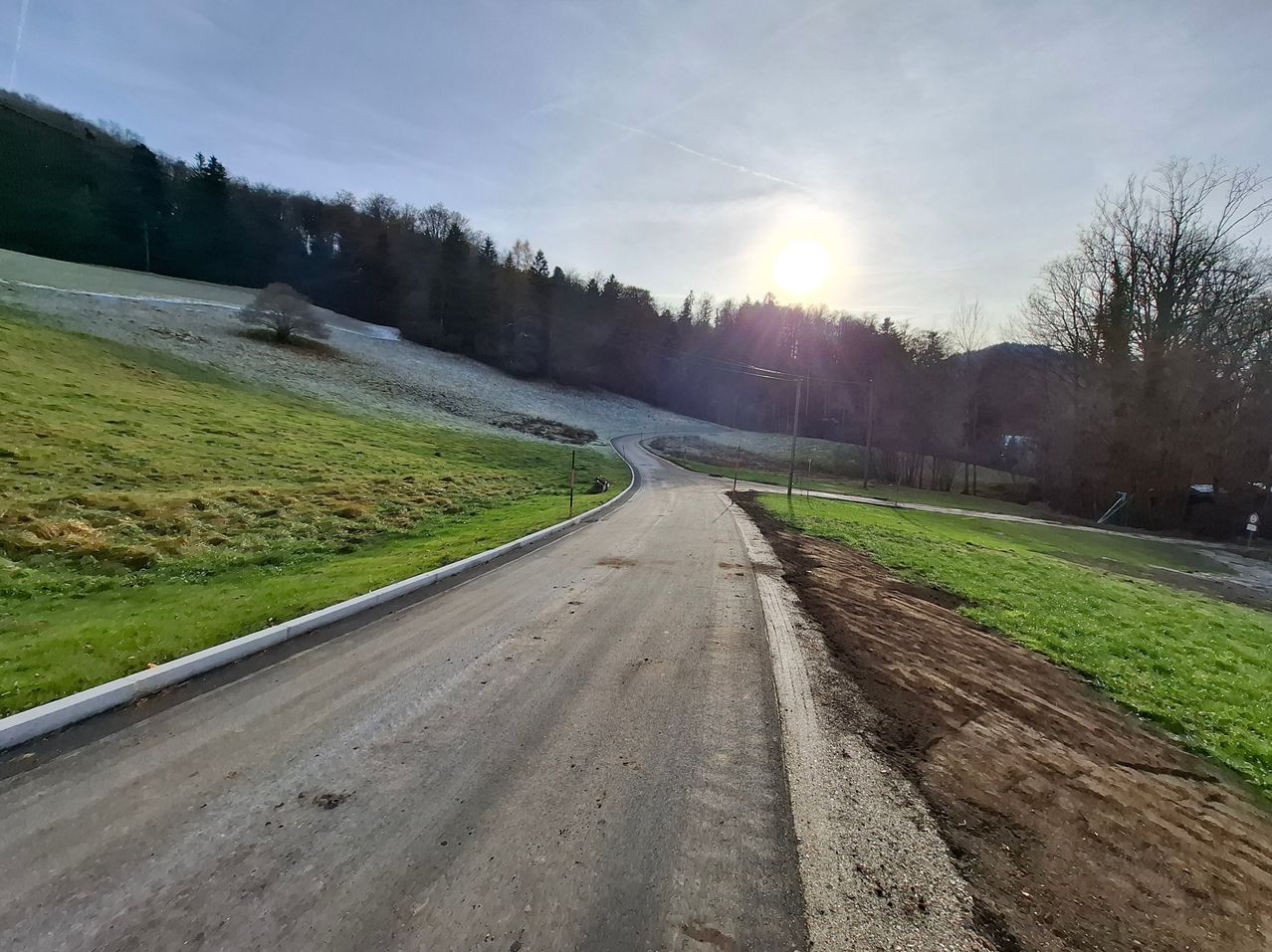 Eine ruhige Landstraße umgeben von üppigen grünen Feldern unter einem hellen Sonnenschein, mit einer sanften Kurve und etwas Bauarbeiten in der Nähe.