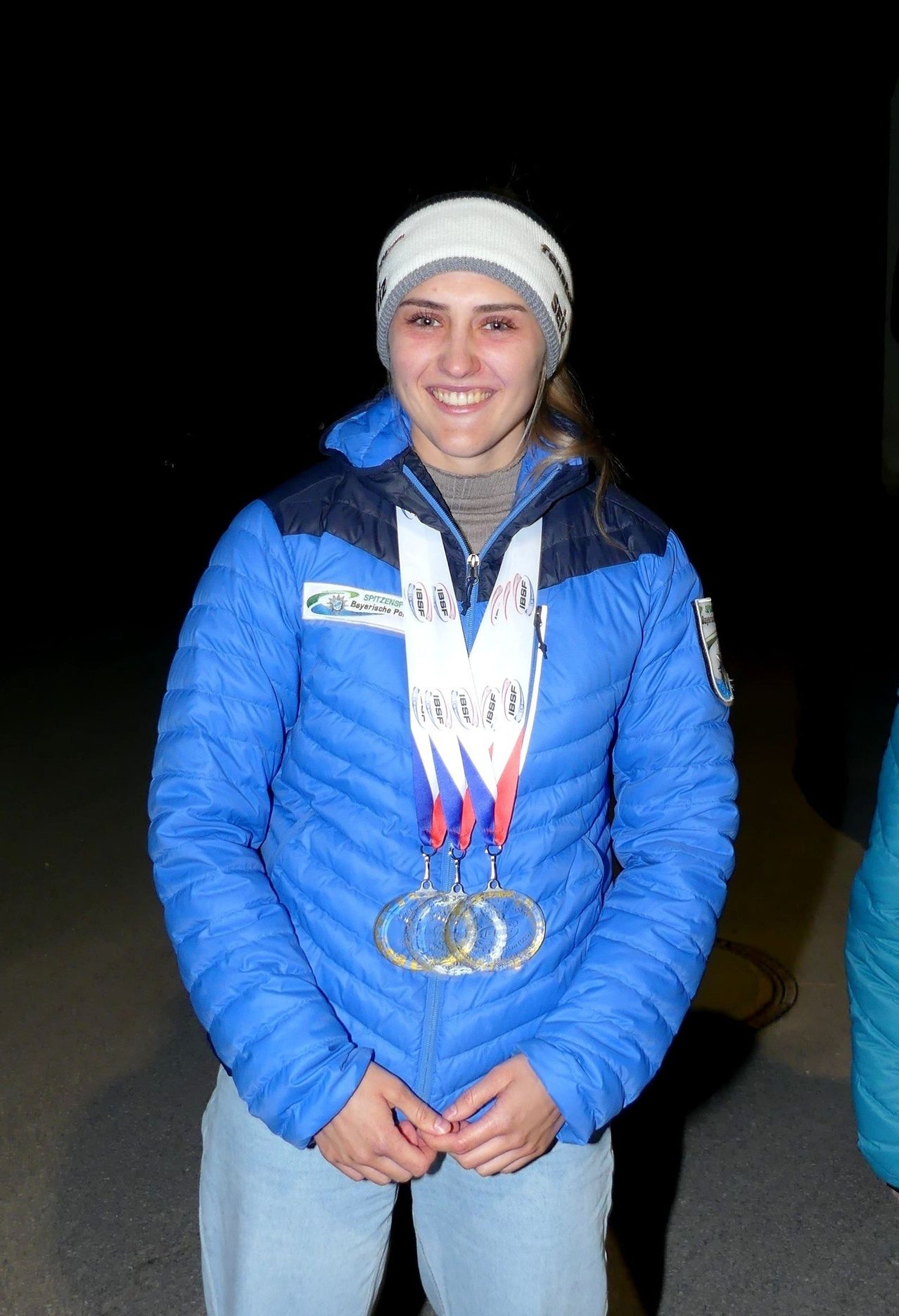 Eine Frau in einer blauen Jacke und einer weißen Mütze posiert mit drei Medaillen um den Hals für ein Foto.