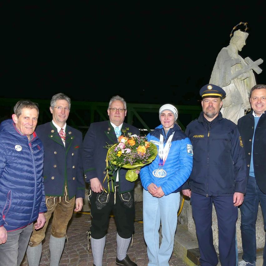 Eine Gruppe von Menschen steht vor einer Statue, eine Frau mit Blumen und Medaillen, zwei Männer in Uniformen und zwei Männer in traditioneller Kleidung.