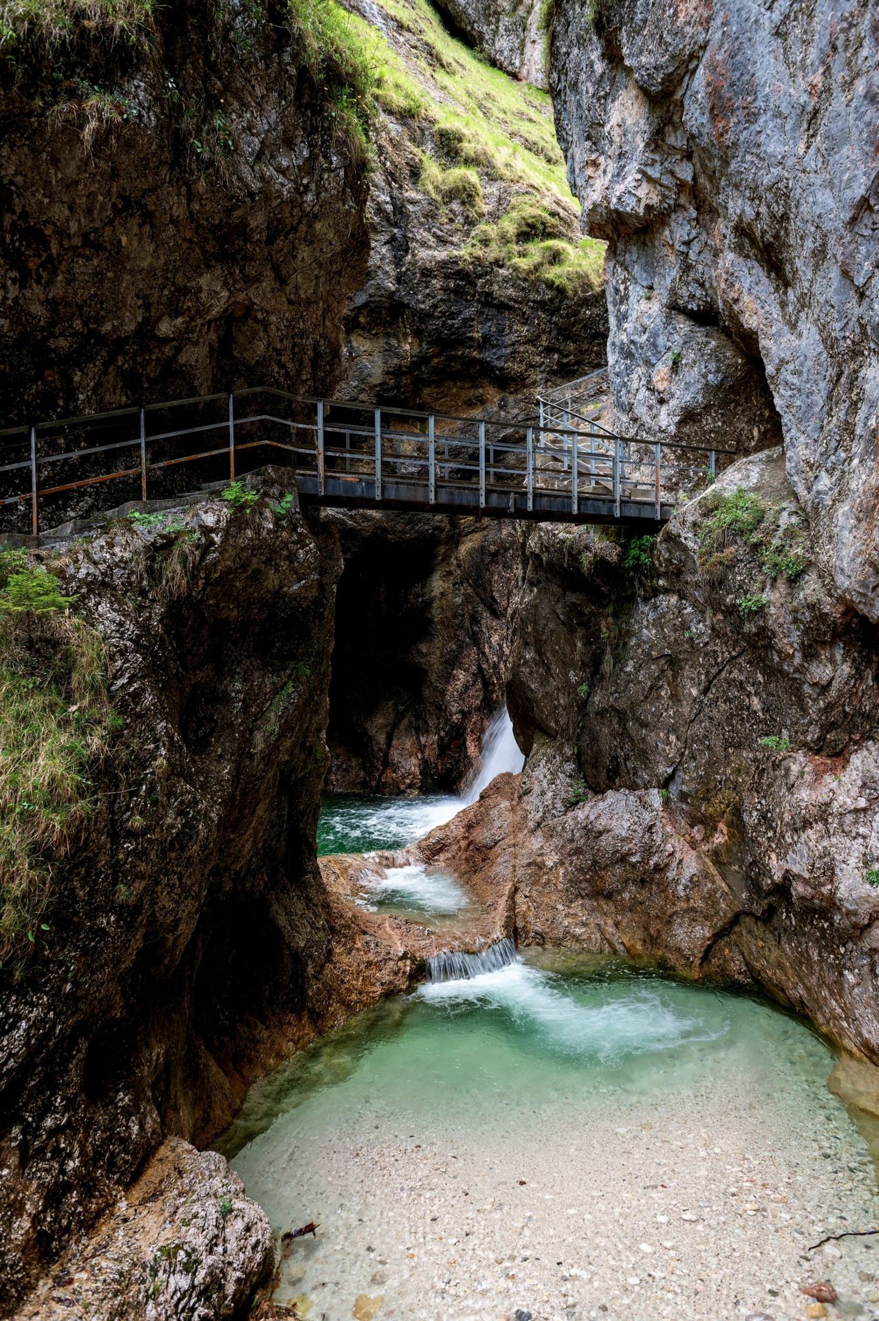 Ein Gebirgsbach fließt durch eine enge Schlucht mit einer Metallbrücke darüber. Die Felswände sind rau mit Felsen und Grün. Wasser stürzt aus einer Höhe und bildet ein kleines Becken darunter.