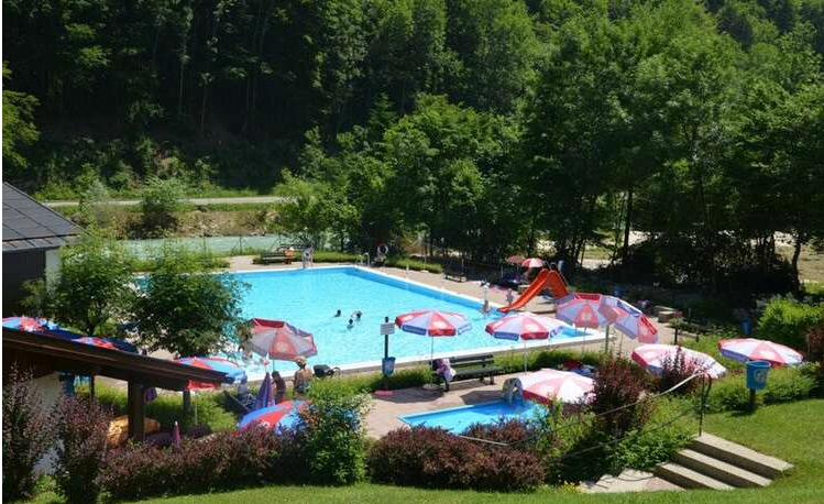 Freibad mit roter Rutsche und Schirmen. Einige Leute schwimmen, andere entspannen. Bäume und Grün umgeben das Gelände.