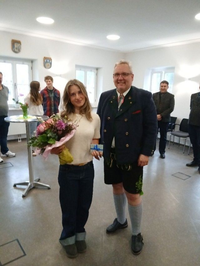 Eine Frau mit Blumen steht neben einem Mann in Uniform. Sie lächeln. Hinter ihnen stehen und sitzen Personen. Im Hintergrund befinden sich Tische, Stühle und Fenster.
