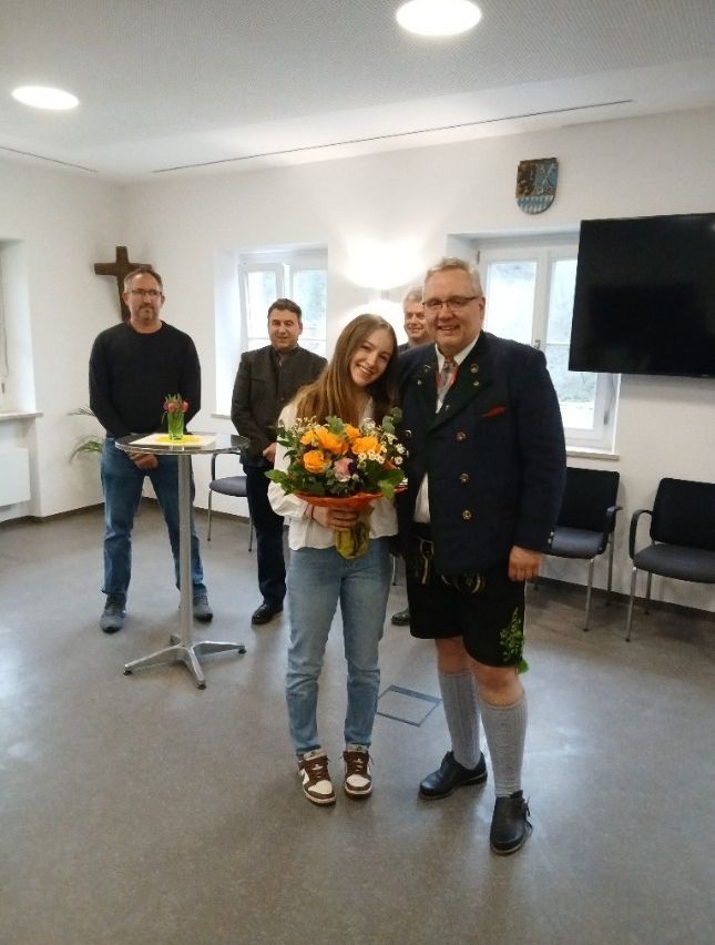 Eine Gruppe von Menschen steht in einem Raum. Ein Mann und eine Frau lächeln und halten einen Blumenstrauß. Sie stehen nebeneinander. Hinter ihnen steht ein Mann. Ein Tisch mit einem Blumenvase steht vor ihnen. Stühle sind dahinter aufgestellt. Ein Kreuz ist an der Wand montiert. Ein Monitor ist an der Wand auf der rechten Seite montiert.