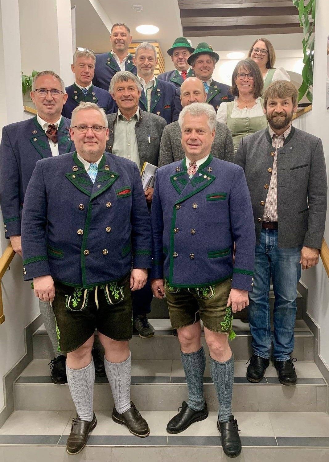 Eine Gruppe von Männern und Frauen in traditioneller Kleidung steht auf einer Treppe. Die Männer tragen passende blaue Jacken mit grünem Besatz und grüne Shorts mit Blattdesigns. Sie posieren für ein Foto.