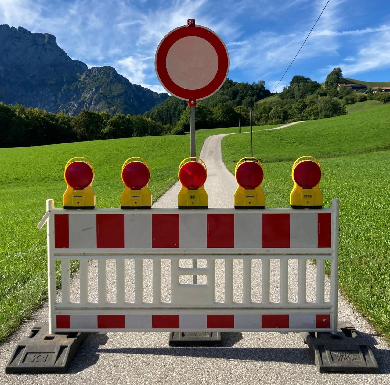Ein Weg mit einer Barriere, die blinkende Lichter und ein Rundum-Zeichen trägt, blockiert die Straße, vor dem Hintergrund von üppigen grünen Feldern und Bergen.