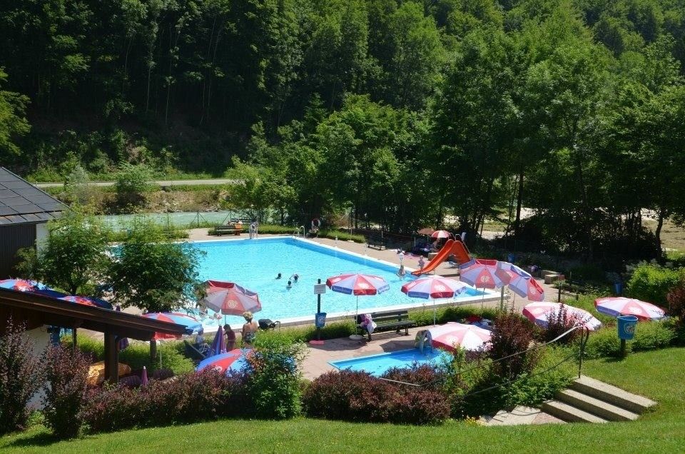 An outdoor swimming pool with several people swimming and others lounging under colorful umbrellas. Trees and greenery surround the area, with a river and mountains in the background.