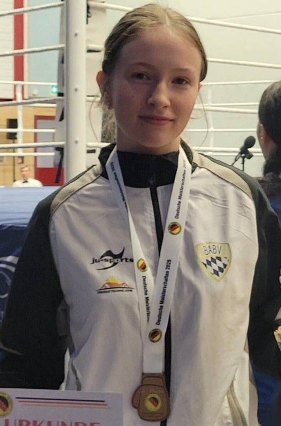 Ein Mädchen in einer weißen und schwarzen Sportjacke hält eine Goldmedaille und lächelt für die Kamera. Sie hat eine weiße Schleife mit deutschem Text um den Hals.