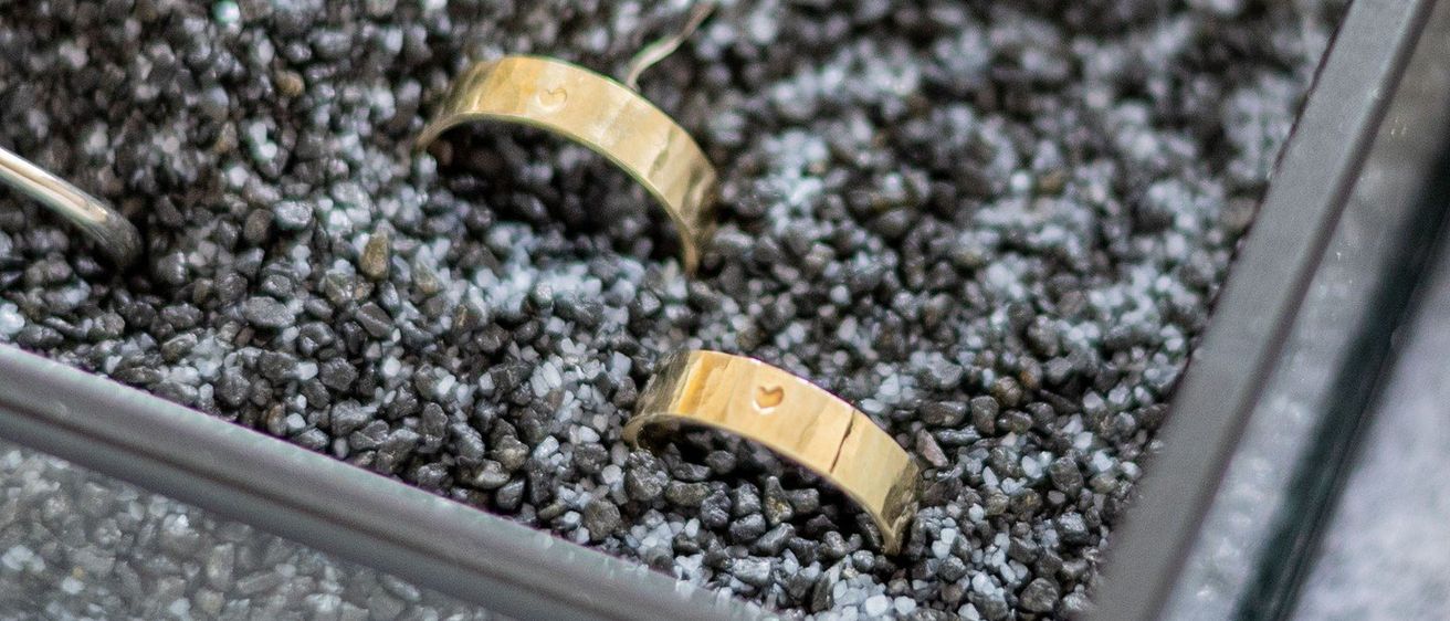 Zwei Goldringe, einer mit Herzdesign, liegen in einer Glasschale mit kleinen Steinen auf einer grauen Oberfläche.