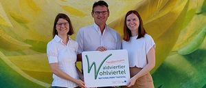Drei Personen stehen vor einer farbenfrohen Wandmalerei und halten ein Schild mit der Aufschrift 'Wohlviertel Nationalpark Thaital.' Die Frau links trägt eine Brille und ein weißes Hemd, der Mann in der Mitte trägt eine Brille und ein weißes Hemd. Die Frau rechts trägt ein weißes Hemd und eine braune Rock.