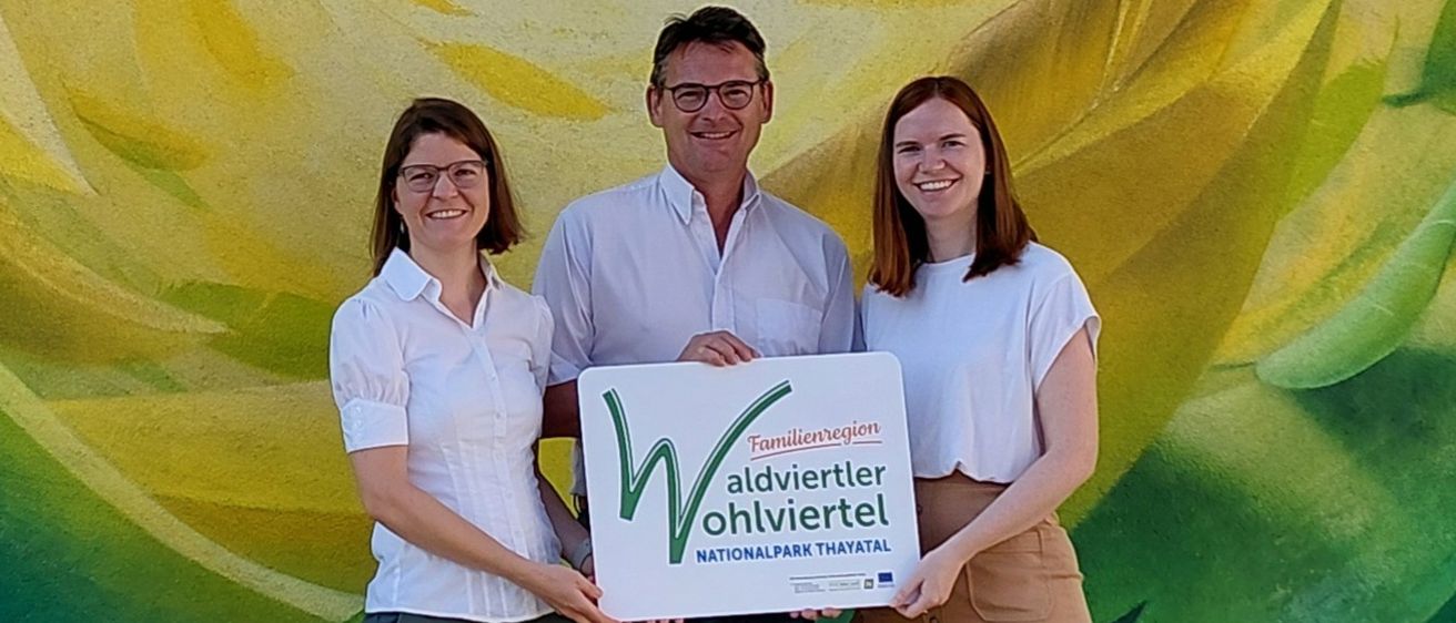 Drei Personen stehen vor einer farbenfrohen Wandmalerei und halten ein Schild mit der Aufschrift 'Wohlviertel Nationalpark Thaital.' Die Frau links trägt eine Brille und ein weißes Hemd, der Mann in der Mitte trägt eine Brille und ein weißes Hemd. Die Frau rechts trägt ein weißes Hemd und eine braune Rock.