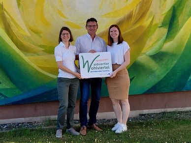 Drei Personen stehen vor einer farbenfrohen Wandmalerei und halten ein Schild mit der Aufschrift 'Wohnviertel' in Grün. Die Frau links trägt eine Brille und ein weißes Hemd, der Mann in der Mitte trägt eine Brille und ein weißes Hemd und die Frau rechts trägt ein weißes Hemd und einen braunen Rock.