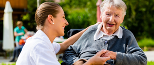 Eine Krankenschwester hält die Hand einer lächelnden älteren Frau im Freien, mit Bäumen und Gebäuden im Hintergrund.