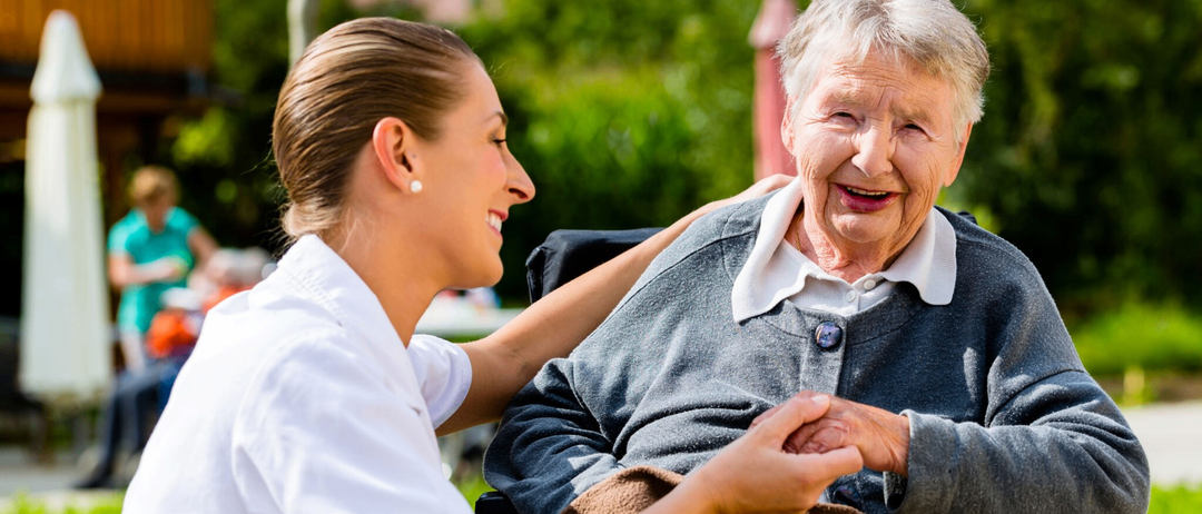 Eine Krankenschwester hält die Hand einer lächelnden älteren Frau im Freien, mit Bäumen und Gebäuden im Hintergrund.