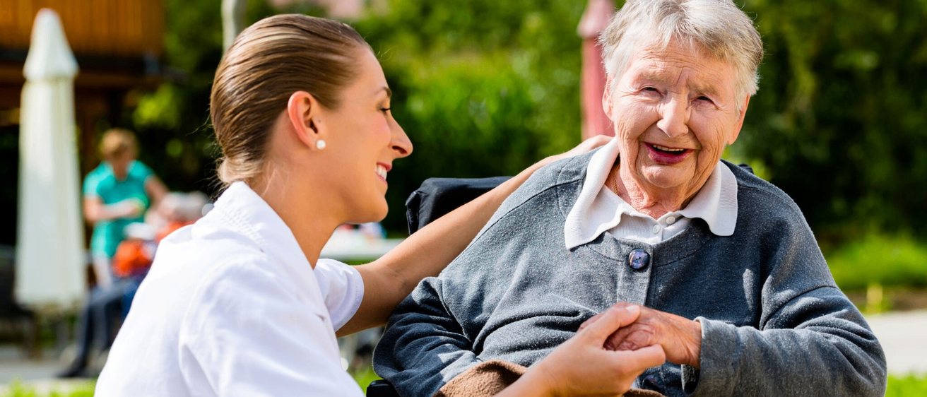 Eine Krankenschwester hält die Hand einer lächelnden älteren Frau im Freien, mit Bäumen und Gebäuden im Hintergrund.