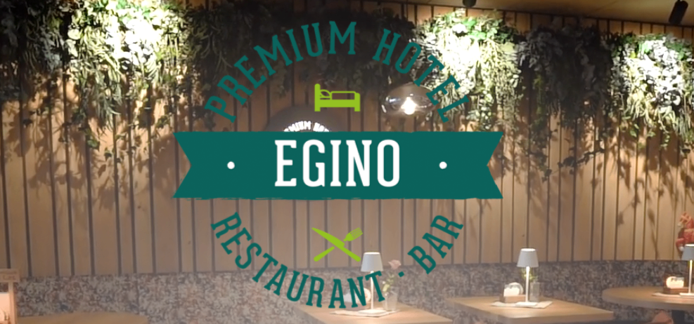 Innenansicht eines Premium-Hotels mit einem Restaurant und einer Bar namens Egino. Die Wände sind mit hängenden Pflanzen und Holzpaneelen geschmückt. Zwei Tische sind mit Lampen und Besteck gedeckt.