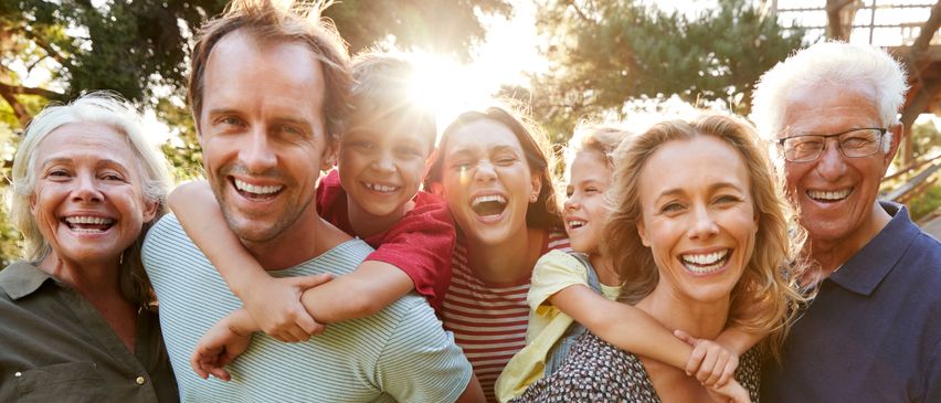 Eine Familie mit fünf Mitgliedern, darunter zwei Erwachsene und drei Kinder, lächelt und posiert für ein Foto in einem Park. Die Sonne scheint hell hinter ihnen.