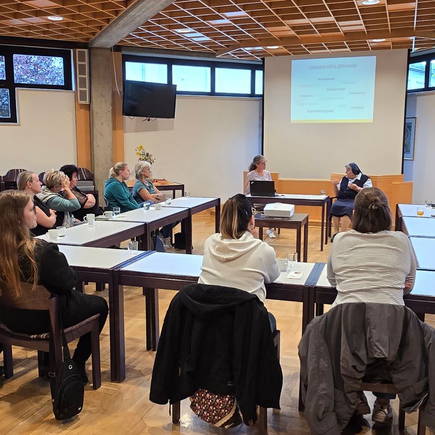 Ein Klassenzimmer mit mehreren Frauen, die an Tischen sitzen. Eine Projektionsleinwand zeigt Inhalte, und eine Frau steht an einem Schreibtisch mit einem Laptop. Eine Frau in einer Nonnentracht sitzt in der Nähe und führt ein Gespräch.