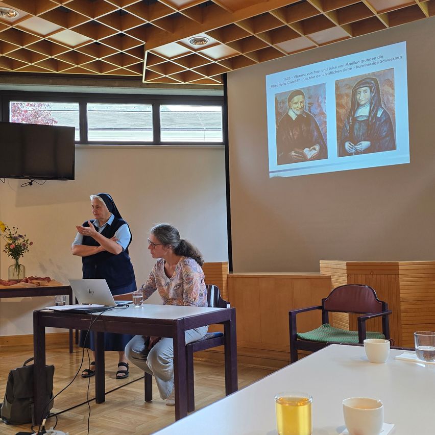 Zwei Frauen sitzen an einem Tisch in einem Raum mit Laptop, einer Vase mit Blumen und einer Projektionsleinwand. Die Frau mit Brille spricht, während die Nonne zuhört.