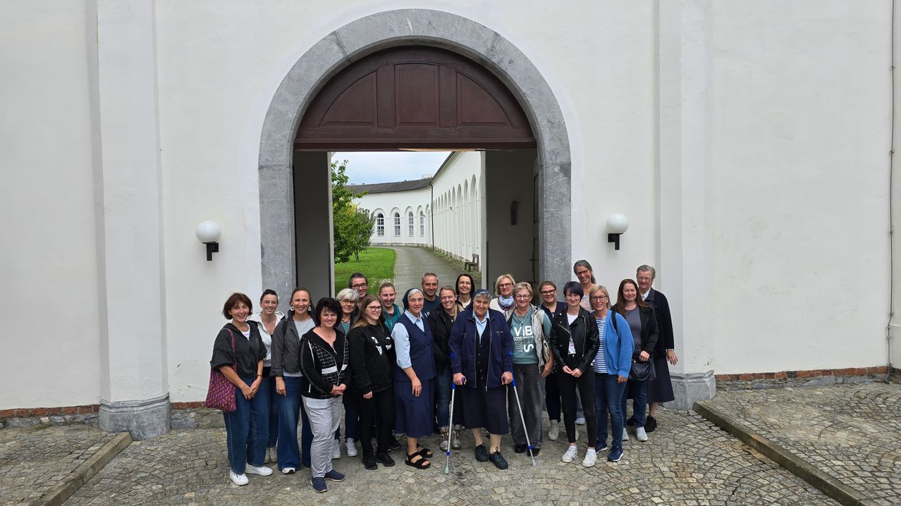 Eine Gruppe von Menschen, darunter Frauen mit Brille und eine Nonne, posiert für ein Foto vor einem weißen Gebäude mit gewölbten Durchgängen.